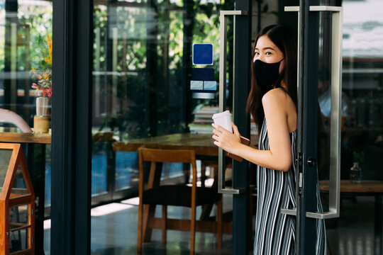 Young Beautiful Asian Woman Wearing A Surgical Face Mask While Leaving A Coffee Cafe Shop. Attractive Female Customer Buying Coffee And Holding A Disposable Coffee Cup. Covid-19 Prevention.