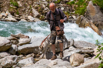 Fototapeta premium An old man and sled dog walk near the river. Alpine landscape. Active leisure pensioner. An elderly man is smiling. Walk with Siberian Husky.
