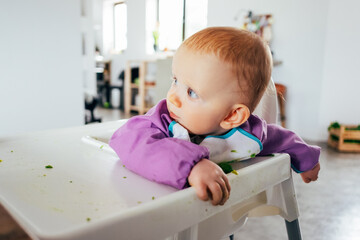 Portrait of curious little child sitting on highchair messy after feeding. Baby girl wearing bib sitting at dining table. Childcare and infancy concept