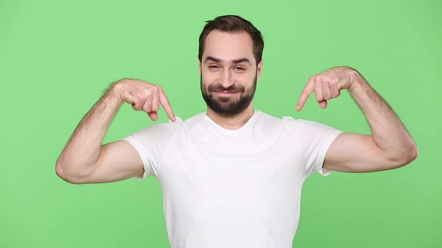 Bearded young guy 20s in white t-shirt isolated on green chroma key background studio. People sincere emotions lifestyle concept. Looking at camera charming smile pointing fingers down on copy space