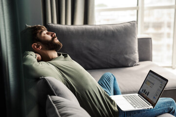 Image of relaxed bearded man resting while working with laptop on sofa