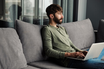 Image of focused young man using laptop while sitting on sofa