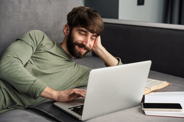 Image of joyful young man using laptop and smiling while lying on sofa