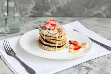 A stack of pancakes with fresh strawberries on a white plate on a light background. A white flower in a glass vase stands next to the Breakfast.