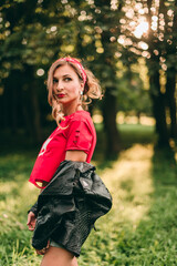 Fototapeta na wymiar Portrait of beautiful young blonde stylish woman with long curly hair and red lipstick in rock style in short denim shorts, red headband, t-shirt, black leather jacket.