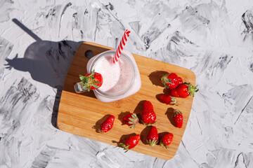 Strawberry smoothie in a glass jar with a red-striped tube. There are strawberries on a wooden Board and a strawberry smoothie next to them. Concept of healthy food. On a light concrete background