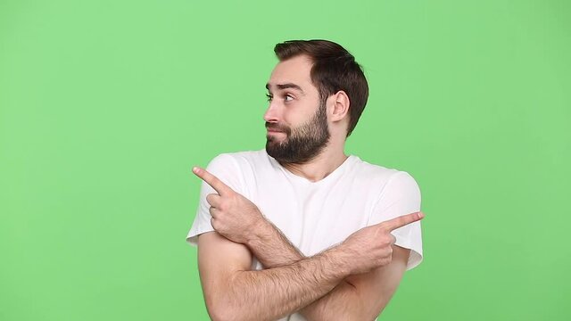 Bearded confused young guy 20s in white t-shirt isolated on green chroma key background studio. People sincere emotions lifestyle concept. Pointing fingers on different sides like says it is not me