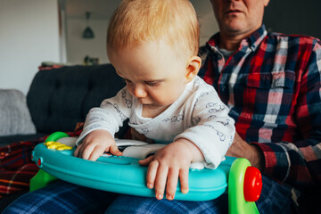 Concentrated baby sitting on fathers lap and playing with toy. Little girl resting at home with grandpa. Childcare and infancy concept