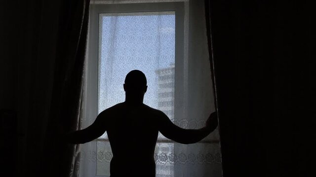Rear view of a man with short hair standing at home and tight-fitting curtains, silhouette against the window, social distance and self-isolation in quarantine isolation.