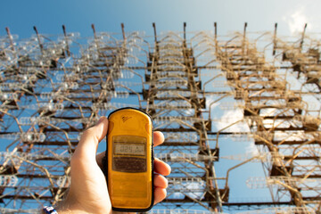 Hand holding a yellow dosimeter indicating radiation on the screen. Enormous Duga radar complex in...