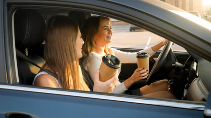 Portrait of two smiling girls driving in car and drinking coffee from paper cups at early morning