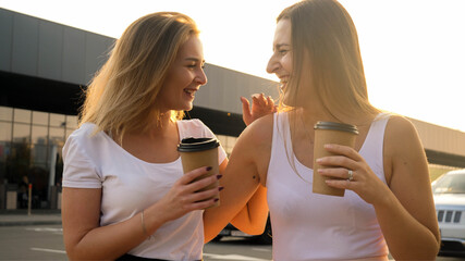 Portrait of two girl friends drinking coffe and laughing on city street at sunset