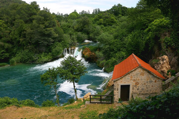 Fototapeta na wymiar Old traditional watermill on river Krka in National Park Krka, Croatia. Beautiful watrefalls in the background. Krka is popular summer travel destination.