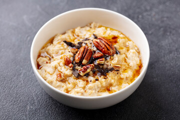 Oatmeal porridge with pecan nuts, maple syrup and chocolate in white bowl. Grey background. Close up.