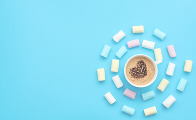 The cup of cappuccino with heart-shaped cocoa on coffee foam and delicious marshmallows on blue background. Happy day, breakfast, good morning concepts. Valentine greeting card. Flat lay style.