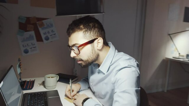 Time lapse 360-degree tracking shot of middle eastern businessman using laptop, talking on phone, drinking coffee, taking notes and reading documents at office desk in the evening