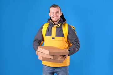 Happy food delivery man holds pizzas in cardboard boxes and holds them out for customer, dressed in yellow uniform and refrigerator bag on his back on blue background. Courier food delivery to home