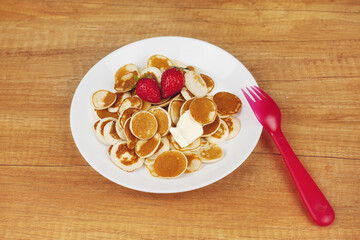 Tiny pancakes are on a white plate with a piece of butter and strawberries on a wooden background, next to a red fork, top view, space for text
