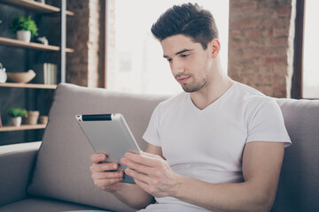 Close-up portrait of his he nice attractive focused funky guy sitting on divan reading ebook using online service hobby at modern industrial loft brick interior style living-room