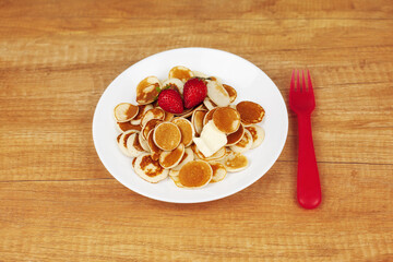 Tiny pancakes are on a white plate with a piece of butter and strawberries on a wooden background, next to a red fork, top view, space for text