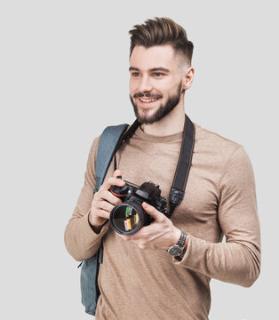 Young cheerful man photographer takes images with digital camera isolated on white background. Studio shot. Travel, photography, professional freelance work, hobby and active lifestyle concept