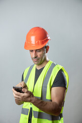 operaio con caschetto protettivo arancione e gillet giallo manda un sms dal suo cellulare, isolato su sfondo grigio