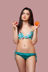 a young beautiful brunette woman in a blue swimsuit with fresh oranges and apples in her hands on a pink isolated background