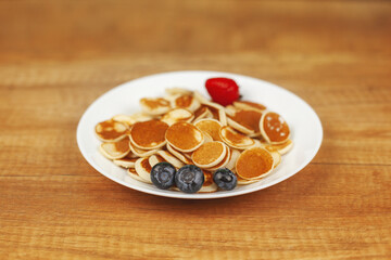 Tiny pancakes sit on a white plate with blueberries and strawberries on a wooden background. Trendy Breakfast, delicious dessert. Space for text