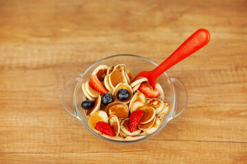 Mini pancakes with strawberries and blueberries in a bowl on a wooden table, top view. Trendy food - pancake cereal