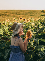 A young blonde woman with short hair in a hat and blue dress stands in a field near yellow ripe sunflowers with seeds in summer at sunset.