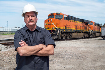 Male worker wearing white construction hard hat with arms folded standing in front of diesel train locomotive in train yard