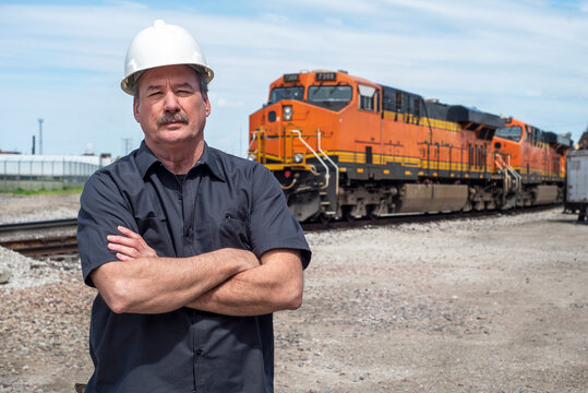 Male Worker Wearing White Construction Hard Hat With Arms Folded Standing In Front Of Diesel Train Locomotive In Train Yard