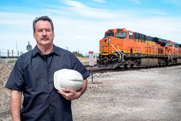 Male worker holding white construction hard hat standing in front of diesel train locomotive in train yard