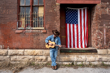 Man wearing cowboy hat, playing acoustic guitar outside next to factory loading dock with American...
