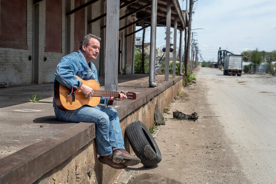 Man Playing Acoustic Guitar Wearing Jeans Outside In The City Sitting On Old Loading Dock 