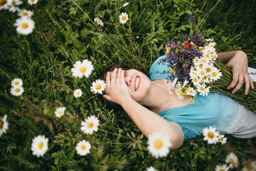 Portrait of a young white beautiful woman in a blue T-shirt and no makeup with a bouquet of fresh wildflowers and and a wreath on her head lying on green grass in the summer nature in the mountains.