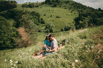 Fototapeta na wymiar Young white happy romantic beautiful couple hugging, sitting on plaid on green grass on nature in summer in mountains. Woman with a wreath of daisies on her head. Prak, nature, rest, love.