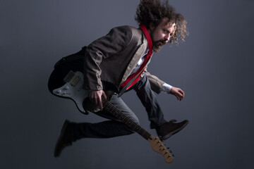 Obraz premium defocused studio portrait of a young bearded, curly-haired guy with long hair, in a jacket, jumping with a guitar, looking at the camera. On a gray background. Dynamic male dramatic portrait