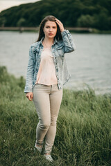 Young white beautiful sad woman in denim, jeans clothes stands on nature in summer near lake at sunset. 