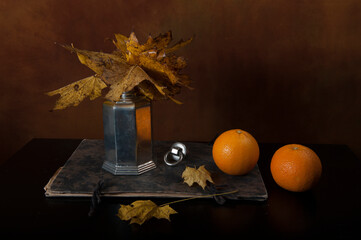 Two oranges, vintage herbarium and tin carafe with autumn leaves. Faded fall foliage in pewter container.