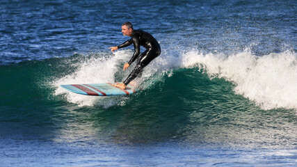 A male somewhat older surfer on a surfboard rides a wave with a lot of spray in front of a Mediterranean beach. The board is red and white and the man has a black surf suit.