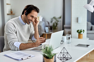 Young happy businessman wearing headset while working on laptop at home.