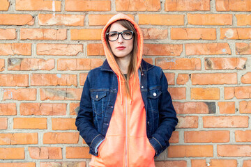 Serious young girl wearing glasses, arms crossed, wearing a hoodie, wearing a hood over his head and a denim jacket on a red brick wall