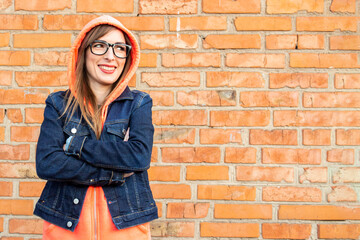 Smiling young woman in a denim jacket and glasses, looking to the side with her arms crossed against a red brick wall