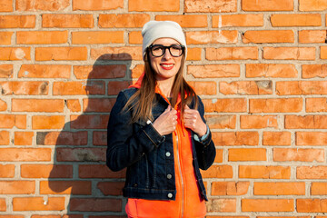 Smiling young girl in glasses, a light hat and in a denim jacket on a background of a red brick wall