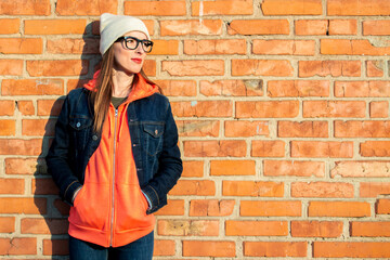 Young girl with glasses, hat and a light denim jacket, holding hands in the blouse and looking to the side on a red brick wall