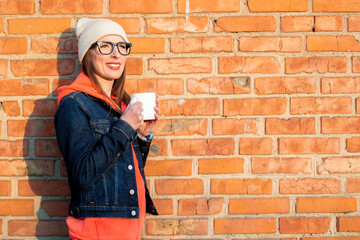 Young smiling woman in glasses holding a white cup on the background of brick wall wearing a gray cap, hoodie and denim jacket. Shot under natural light