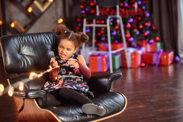 Beautiful kid girl 5-6 year old wearing stylish dress sitting in armchair over Christmas tree in room. Looking at camera. Holiday season.