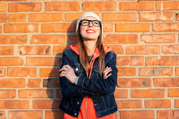 Young woman arms crossed looking to the top of the glasses on the background of a brick wall, wearing a gray cap, hoodie and denim jacket