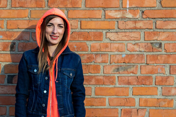 Young woman wearing a hood, in a denim jacket on a background of a red brick wall